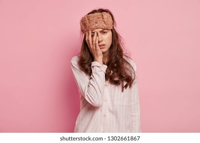 Upset Tired Young Woman Has Not Enough Sleep, Touches Face With Hand, Wears Eyemask And Casual Jumper, Isolated Over Pink Background, Slept Bad At Night. People, Tiredness And Awakening Concept