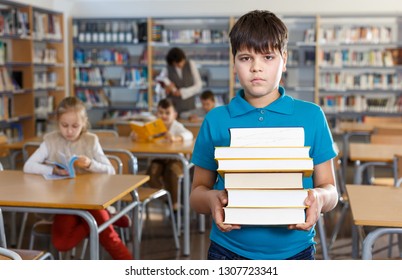 Upset Tween Boy Standing In School Library With Pile Of Books In Hands

