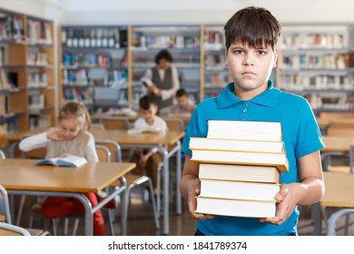 Upset Tween Boy Standing In School Library With Pile Of Books In Hands..