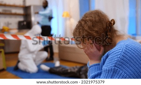 Upset victim wife sitting on couch and covering face with hands. Close up side view of frustrated woman witness sitting at crime scene with policemen investigating on background