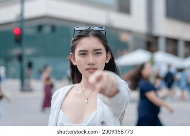 An upset woman calls out a person in public she holds a grudge against. Pointing with her finger towards the camera while looking irate. Outdoor urban city background. - Shutterstock ID 2309358773