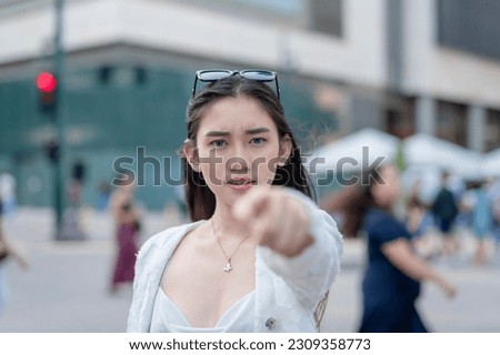 An upset woman calls out a person in public she holds a grudge against. Pointing with her finger towards the camera while looking irate. Outdoor urban city background.