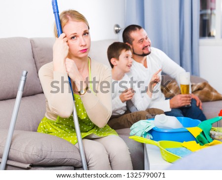 Upset woman with cleaning equipment on couch while her family watching tv
