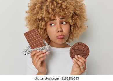 Upset Woman With Curly Blonde Hair Holds Bar Of Chocolate And Cookie Looks Sadly Feels Unhappy Dressed In Casual Jumper Has Sweet Tooth Sugar Temptation Isolated Over White Background. Harmful Food