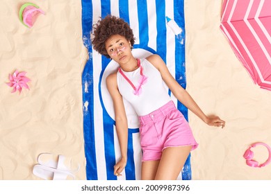 Upset Woman With Curly Hair Sad To Get Sunburn After Sunbathing All Day At Beach Applies Sunblock Lotion Dressed In Summer Clothes Lies On Striped Towel Poses At Seaside Different Items Around