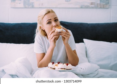 upset woman in pajamas looking at camera while eating cake in bed alone