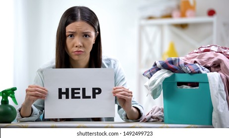 Upset woman showing help word on cardboard, laundry basket with clothes on table