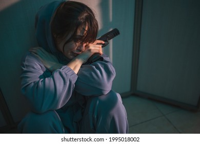 Upset Woman Sitting On The Floor In The Dark, Holding A Gun And Crying After Committing Murder In Self-defense; Scared Woman Victim Of Domestic Violence Holding A Gun For Self-defense