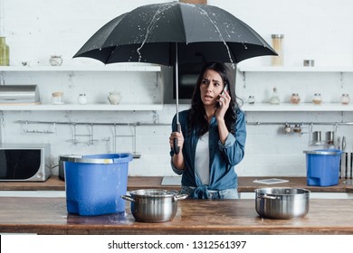 Upset woman standing under umbrella in kitchen and calling plumber