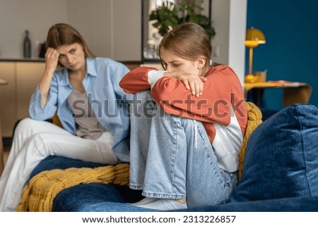 Upset worried mother looking at crying moody teenage daughter, sitting together on sofa, upset frustrated mom and teen girl child not talking after argument, having communication problems 