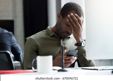 Upset Young African Man Reading Message On His Mobile Phone While Sitting At His Desk