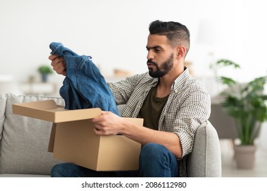 Upset Young Arab Guy Unboxing Carton Package, Receiving New Clothes, Unhappy About Delivered Item At Home. Online Shopping Mistake, Awful Purchase, Wrong Delivery Concept