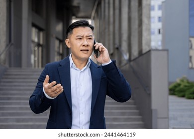 Upset young asian man in business suit standing near building outside and frustrated talking on mobile phone, gesturing with hands. - Powered by Shutterstock