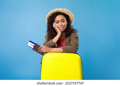 Upset young black woman holding passport and plane tickets, leaning on suitcase, missing vacation because of coronavirus lockdown on blue studio background. Unhappy lady being late for her flight