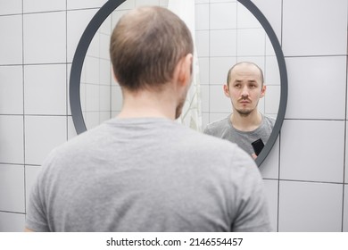 An upset young caucasian man with hair loss problem critically looking at mirror in the bathroom holding hairclipper - Powered by Shutterstock