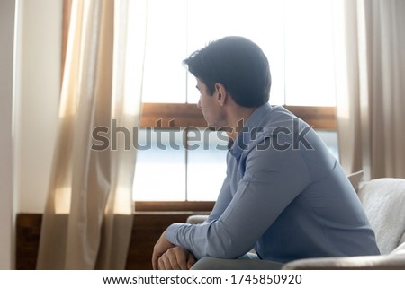 Upset young Caucasian man sit on couch in living room look in distance thinking or pondering, unhappy sad millennial male feel lonely lost in thoughts at home, outcast or loner, solitude concept