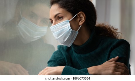 Upset young Caucasian woman in medical facial mask look in window feel lonely distressed sick with covid-19. Unhappy female in facemask suffer from coronavirus. Quarantine, corona pandemic concept.