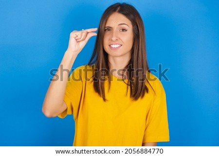 Upset Young european brunette woman wearing yellow T-shirt on blue background shapes little gesture with hand demonstrates something very tiny small size. Not very much