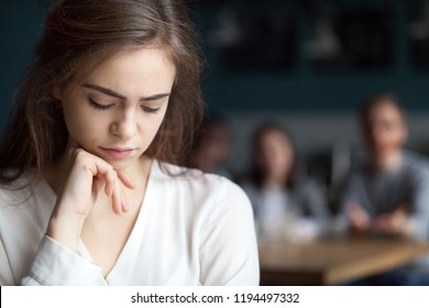 Upset young girl sitting alone in café offended at friends joke, sad millennial woman suffer from low self-esteem, depressed female outcast feeling outsider hanging out with company in coffeeshop