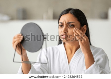 Upset young indian woman in white bathrobe looking at mirror and touching her cheeks, having oily or dry skin problem, puffy eyes in the morning, bathroom interior, closeup photo