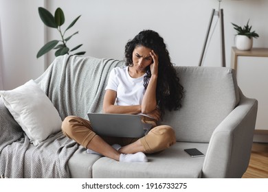 Upset Young Jobless Woman Sitting On Couch With Laptop On Her Lap, Touching Her Head And Looking At Screen, Home Interior. Thoughtful Curly Dark-haired Lady Having Financial Difficulties, Copy Space