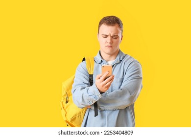 Upset young man with backpack using mobile phone on yellow background