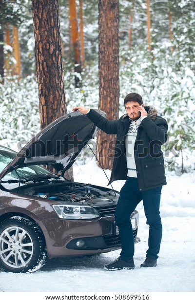 Upset young\
man calling the car service standing on the broken car in the\
winter in the woods. roadside assistance\
car