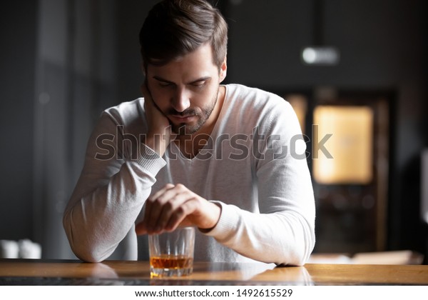 Upset young man drinker alcoholic sitting at bar\
counter with glass drinking whiskey alone, sad depressed addicted\
drunk guy having problem suffer from alcohol addiction abuse,\
alcoholism concept