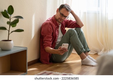 Upset young man holding smartphone waiting for ex-girlfriend call or sms, sitting on floor at home. Unhappy guy looking at phone screen, reading message with bad news. Mental health during break up