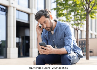 An upset young man sits on a bench on a city street and looks disappointedly at the screen of the phone he is holding in his hands. - Powered by Shutterstock