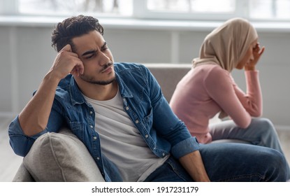Upset young man sitting by his crying wife woman in hijab, having fight at home. Muslim couple suffering from difficulties in relationships, sitting separated on couch in living room