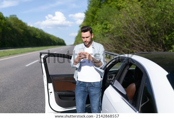 Upset young man standing near car, looking at\
smartphone screen, having problem with mobile connection, trying to\
find network on highway. Road emergencies, calling breakdown\
service, no signal