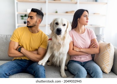 Upset young multinational couple having fight, looking in opposite directions, cute dog sitting on couch between them, indoors. Relationship crisis, marriage conflict, family problems concept