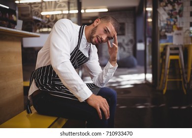 Upset Young Waiter Sitting With Headache While Looking Away At Coffee Shop