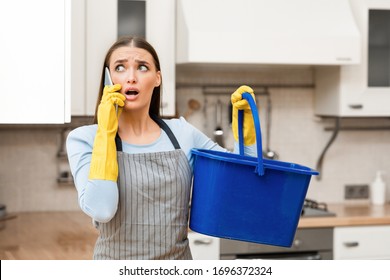 Upset Young Woman Calling Plumber To Fix Leakage In Kitchen, Holding Blue Bucket