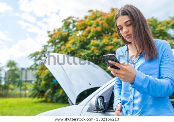 Upset young woman
with cell phone near broken car . Young woman using mobile phone
while looking at broken down car on street. Young woman with broken
car calling for help 