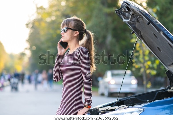 Upset young woman driver talking angrily on cell\
phone with assistance service near a broken car with open hood on a\
city street.