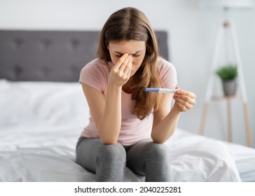 Upset Young Woman Holding Pregnancy Test, Touching Her Face In Stress On Bed At Home. Depressed Millennial Lady Being Unexpectedly Pregnant Or Unable To Have Baby, Having Maternity Problem