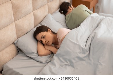 Upset young woman lies in bed turned away from her partner, capturing moment of emotional distance and tension in relationship within bedroom setting - Powered by Shutterstock