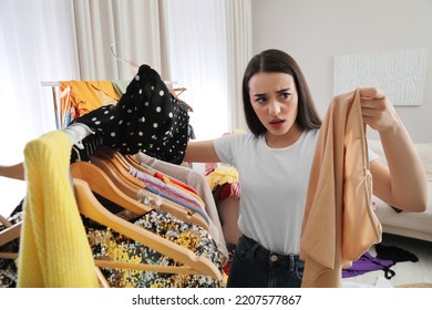 Upset Young Woman Looking Through Clothes On Rack In Room. Fast Fashion