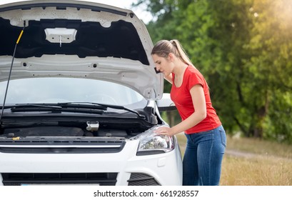 Upset Young Woman Looking Under The Bonnet Of Overheated Car At Countryside