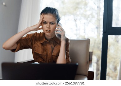 Upset young woman on a phone call, sitting at a desk with a laptop, expressing stress due to unexpected work issues in a cozy, modern environment - Powered by Shutterstock