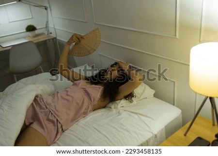 Upset young woman in pajamas feeling hot and using a fan during the summer in bed and having sleeping problems 