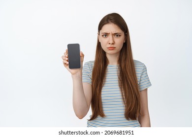 Upset Young Woman Showing Her Phone Screen And Frowning Disappointed. Girl Holds Smartphone, Shows Mobile Screen With Sad Angry Face, Stands Against White Background