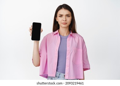 Upset Young Woman Showing Mobile Phone Screen And Frowning, Dislike Application, Having Concerns About Smartphone App, White Background