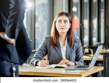 Upsetting Face Expression Of Young Female Employee Sitting And Writing Note On Table With Laptop In Front Of Angry Manager