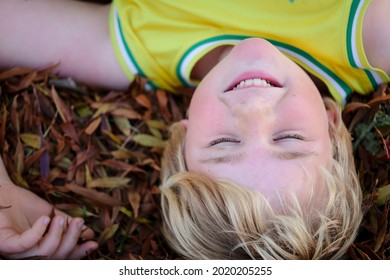 Upside Down Image Of Little Boy Lying In Fallen Autumn Leaves With Eyes Closed And Big Smile