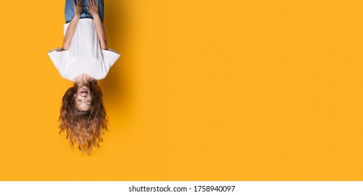 Upside down photo of a caucasian woman in white shirt and jeans smiling on a yellow studio wall with free space