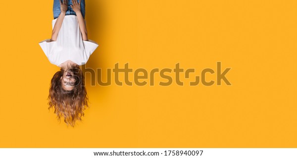 Upside down photo of\
a caucasian woman in white shirt and jeans smiling on a yellow\
studio wall with free\
space
