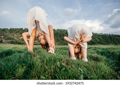 Upside Down Photo Of Happy Children In White Dresses Play On The Grass And Smile In Summer. Funny Portrait Of Two Sisters. Children's Friendship